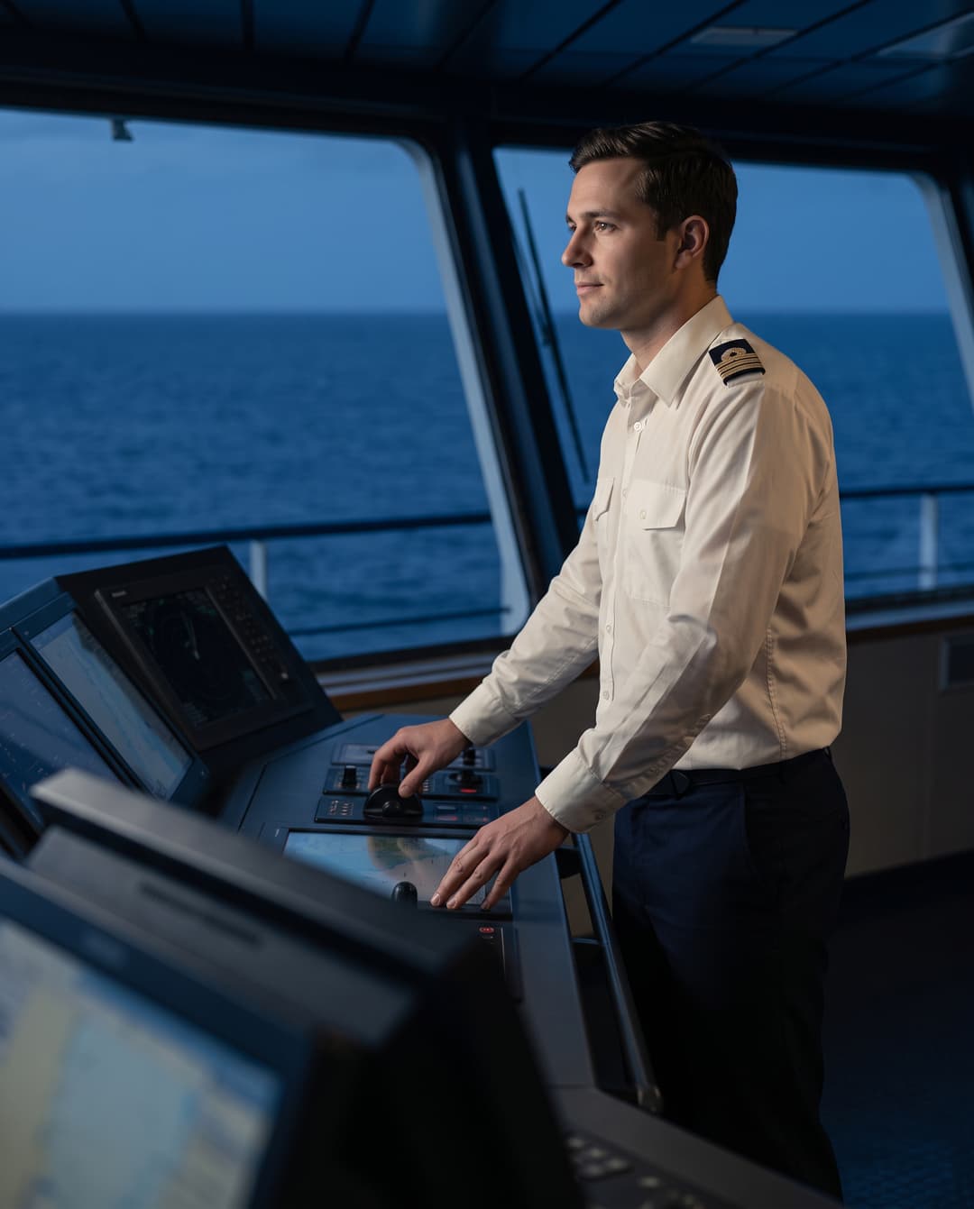 An officer on the bridge with a hand on the controls, the sea behind him